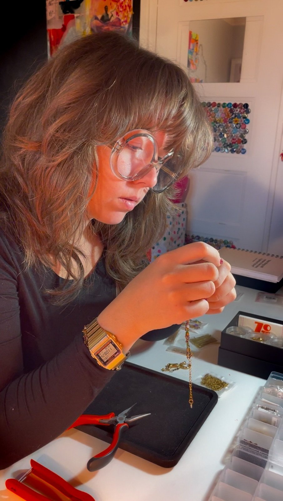 Moi dans mon atelier, lunettes rondes sur le nez, examinant entre mes doigts une chaîne fine ; devant moi un plateau noir, des pinces rouges et mes boîtes de perles