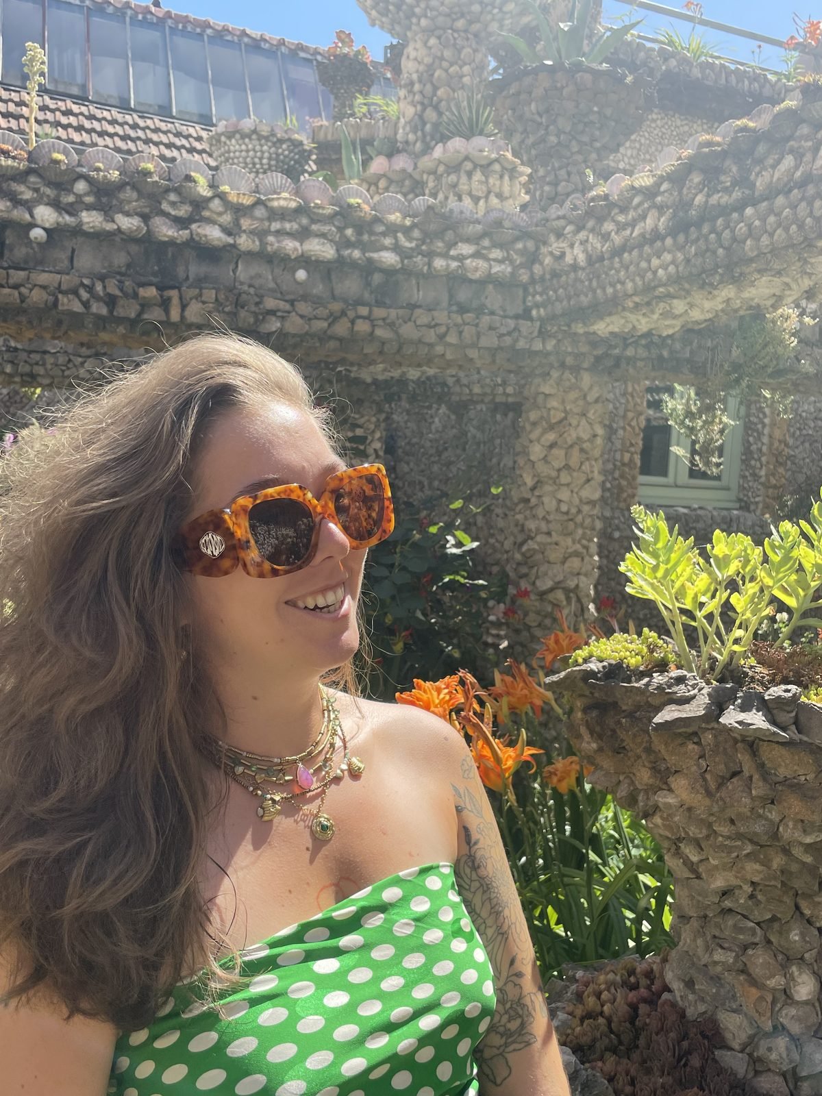 Moi en plein soleil, lunettes écaille et robe verte à pois, plusieurs colliers fins superposés à chaînes dorées et pendentifs en pierre, devant un mur couvert de coquillages et de plantes grasses